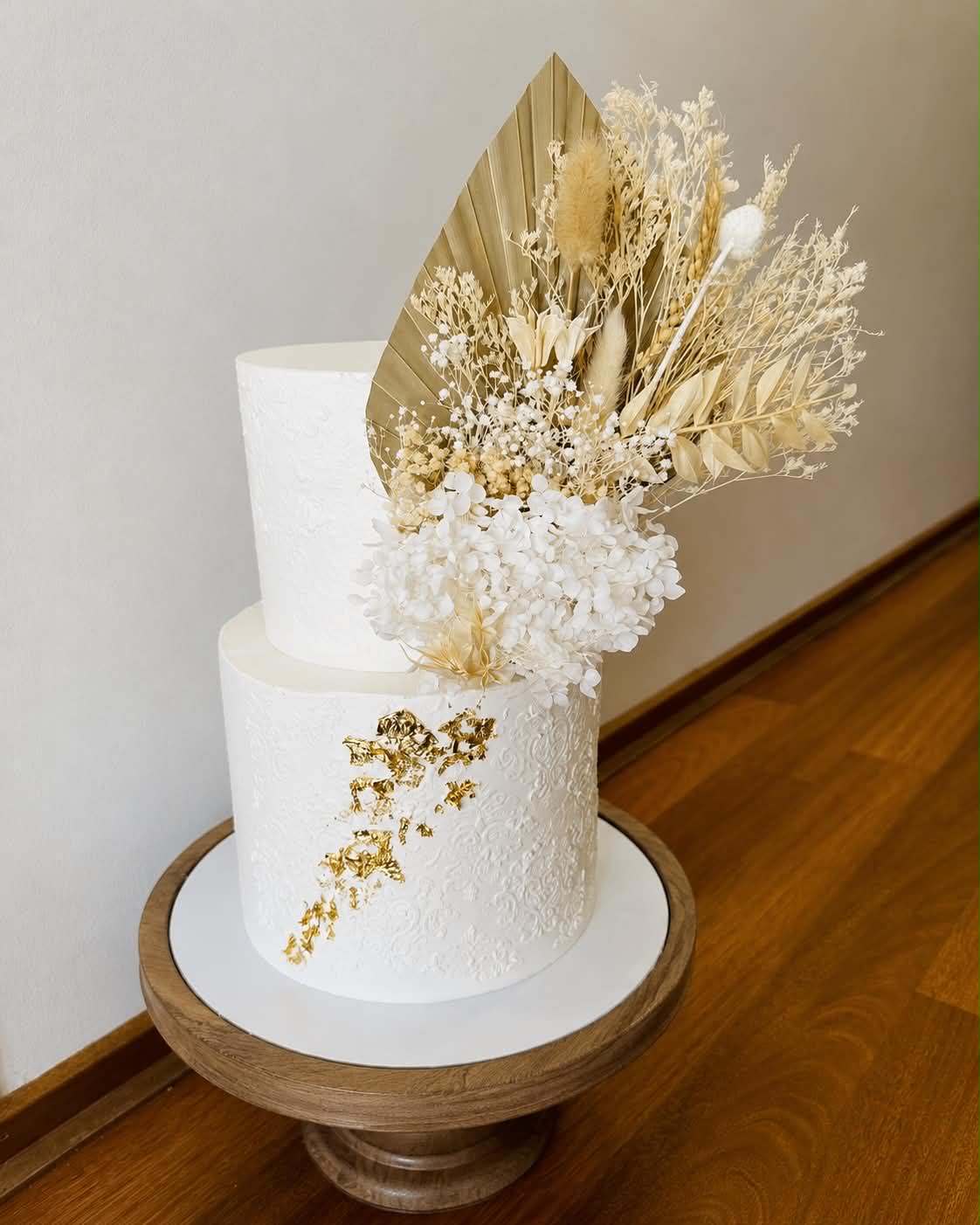 Two tier white fondant wedding cake with lace texture, gold leaf accents and a large dried floral arrangement including pampas grass, bunny tails, baby breath and gold palm leaf fan on top